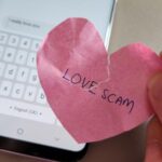 A off focus hand with phone showing message from a scammer and a hand holding a crumpled heart with the word Love Scam written on