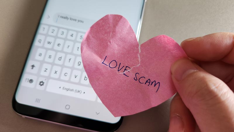 A off focus hand with phone showing message from a scammer and a hand holding a crumpled heart with the word Love Scam written on