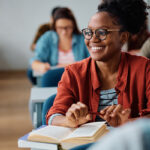 A smiling student in glasses and a red shirt sits in a classroom, holding a pen and book. Others in the background, creating a focused and happy learning atmosphere.
