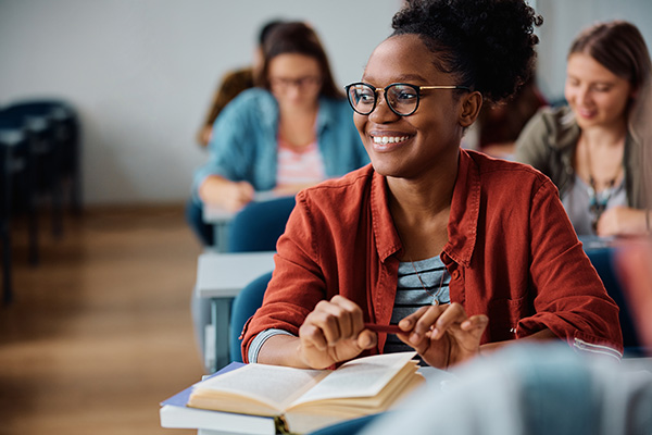 A smiling student in glasses and a red shirt sits in a classroom, holding a pen and book. Others in the background, creating a focused and happy learning atmosphere.