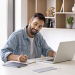 A man is writing on a paper with his laptop on the desk.