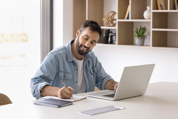 A man is writing on a paper with his laptop on the desk.