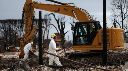 Two workers in protective suits and helmets inspect debris near an excavator in a devastated area, conveying a scene of recovery after destruction.
