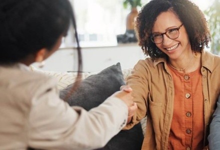 A woman with curly hair and glasses smiles warmly as she shakes hands with another person. They sit on a sofa, creating a friendly atmosphere.